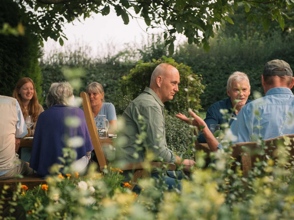 Gruppe von Menschen unterschiedlichen Alters sitzt im Freien um einen Tisch herum und unterhält sich in einem Garten