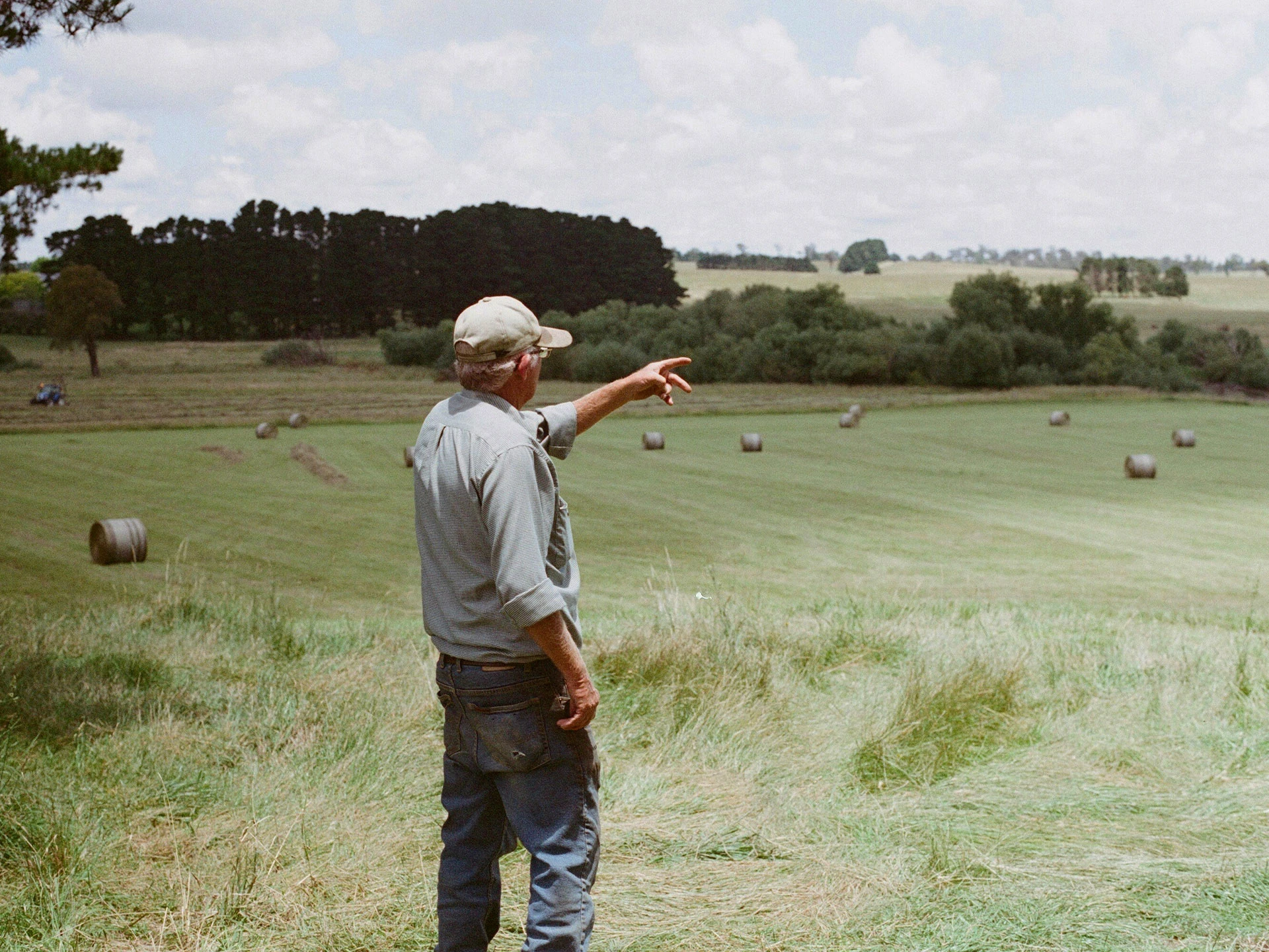 Älterer Mann in Arbeitskleidung zeigt auf ein Feld mit Heuballen