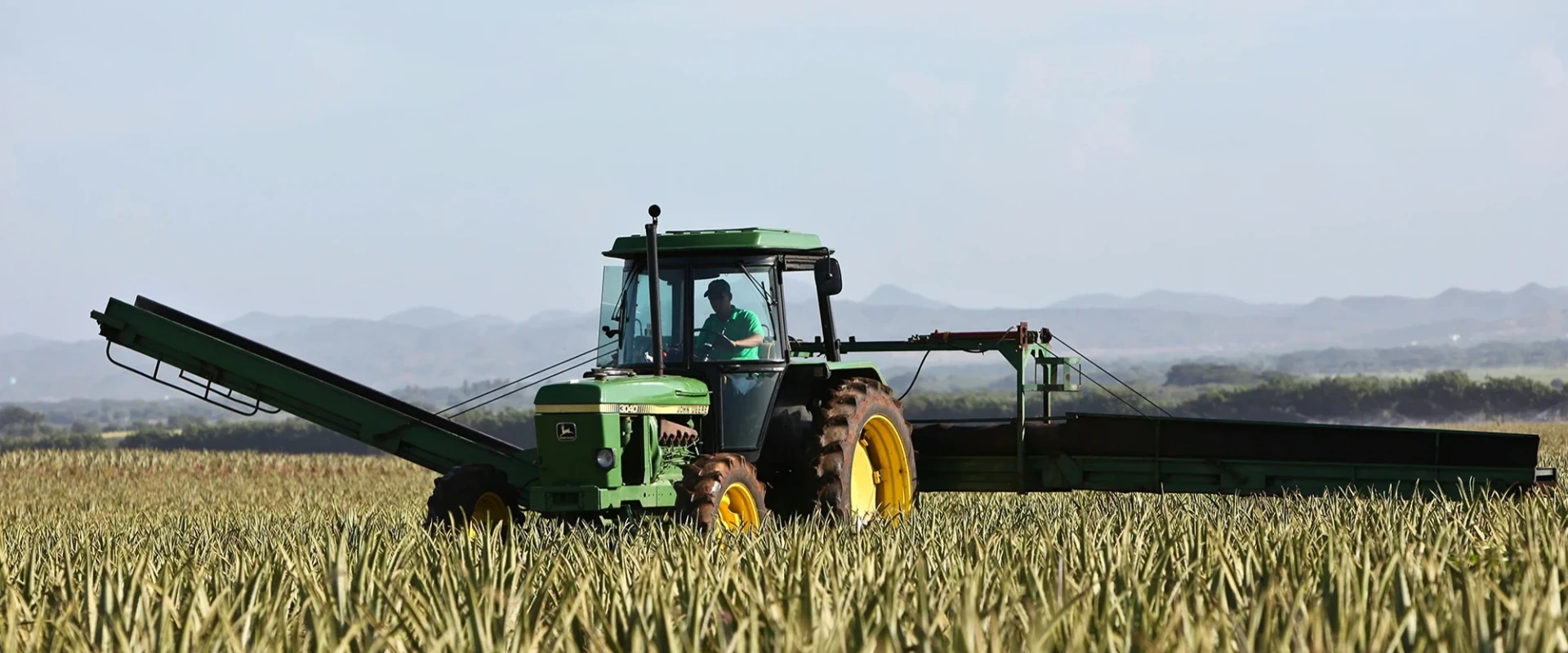 Green tractor on a field during harvest, with mountains in the background