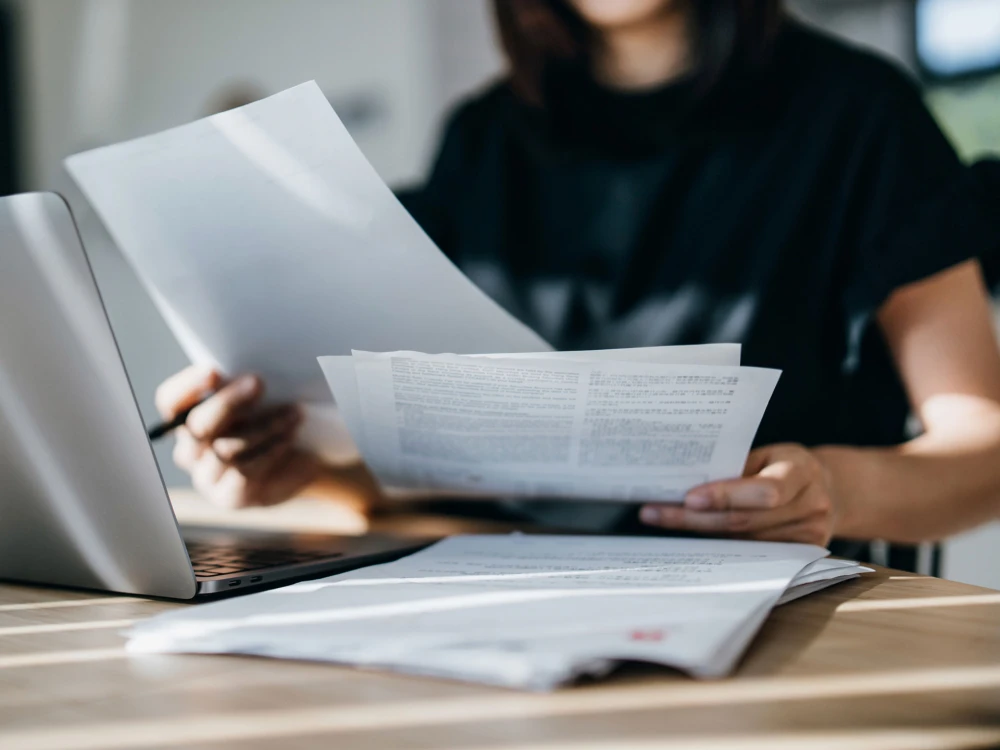 Person reading documents and working on a laptop at a wooden table