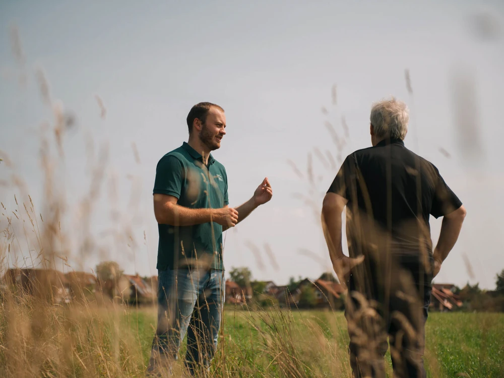 Zwei Männer unterhalten sich auf einer Wiese, einer davon zeigt auf etwas, während er erklärt