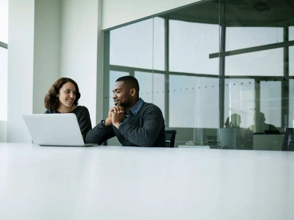 Two business professionals, a woman and a man, sitting at a table in an office and looking at a laptop