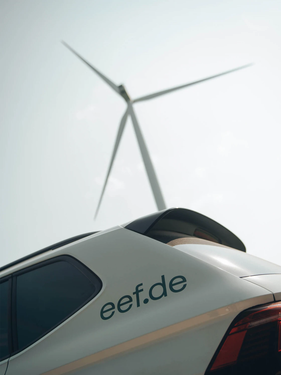 Ein silberfarbenes Auto mit der Aufschrift 'eef.de' auf der Seite, vor einem unscharfen Windrad im Hintergrund