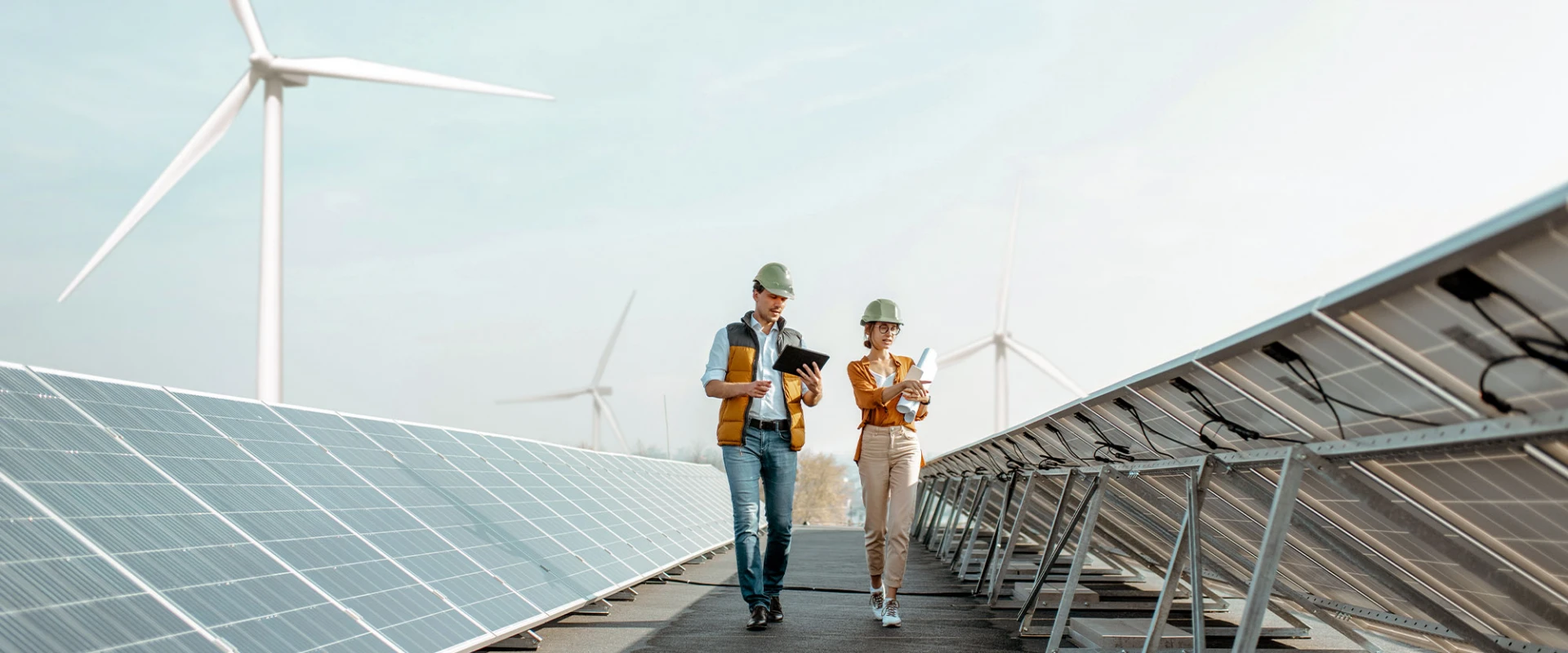 Zwei Ingenieure mit Schutzhelmen gehen zwischen Solarpaneelen und diskutieren, im Hintergrund sind Windräder zu sehen