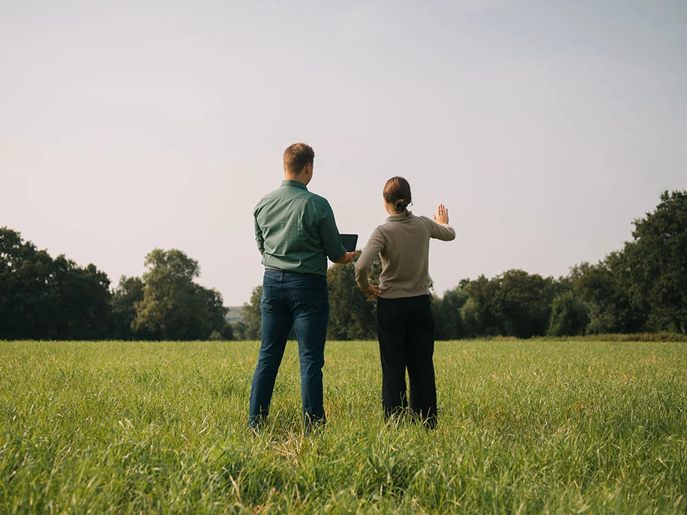 Zwei Personen stehen auf einer Wiese und schauen in die Ferne, eine hebt die Hand, als würde sie etwas zeigen
