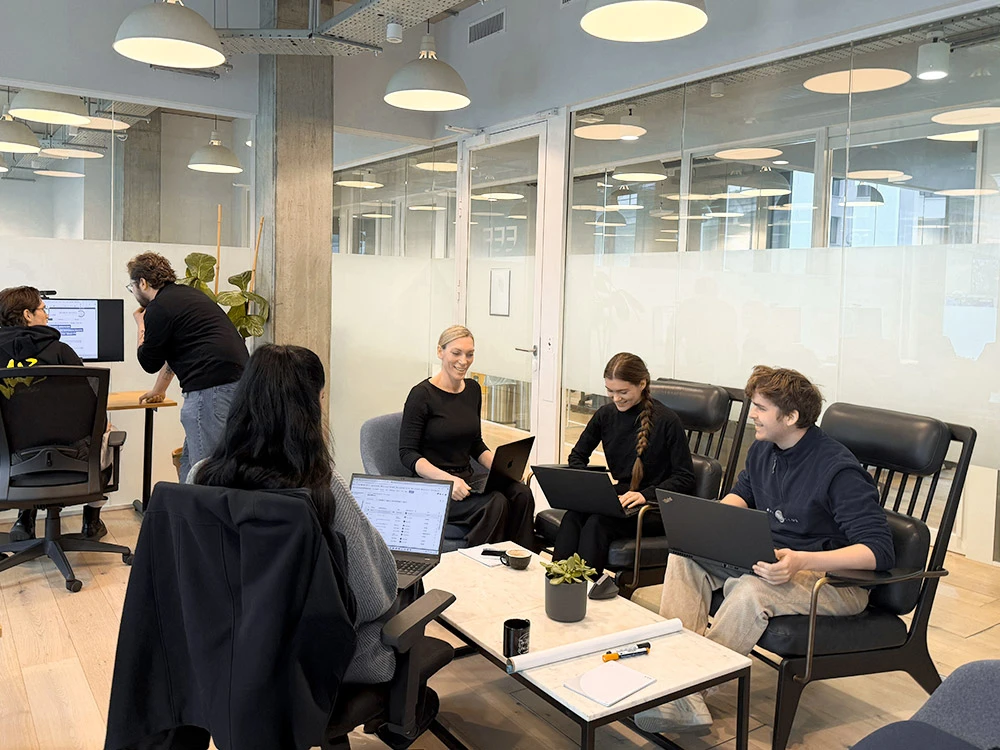 Mehrere Personen, die in einem modernen Büro mit großen Fenstern und Pflanzen an Laptops arbeiten und diskutieren