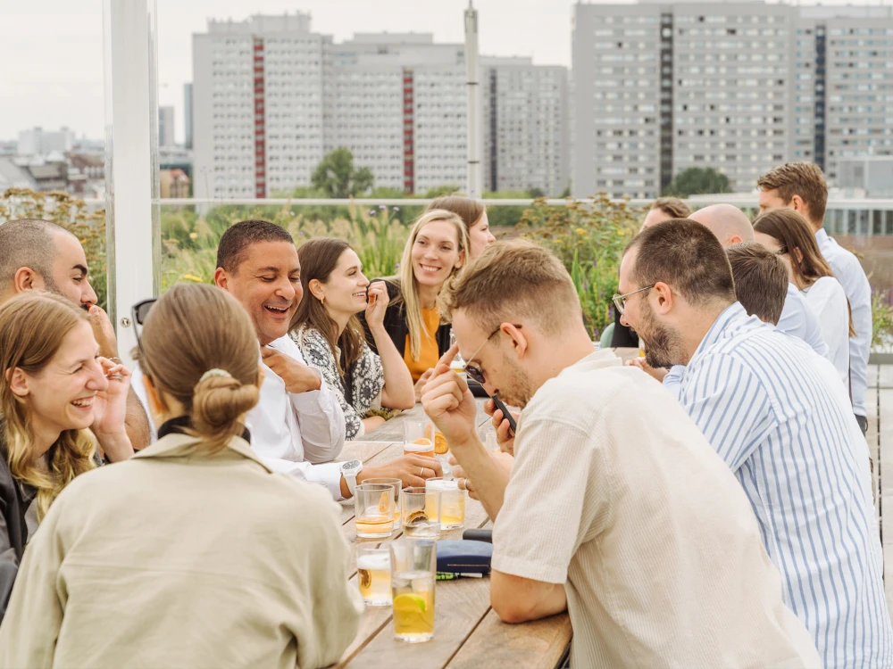 Gruppe von Menschen verschiedener Ethnien lacht und unterhält sich an einem Tisch im Freien mit Getränken vor städtischer Kulisse