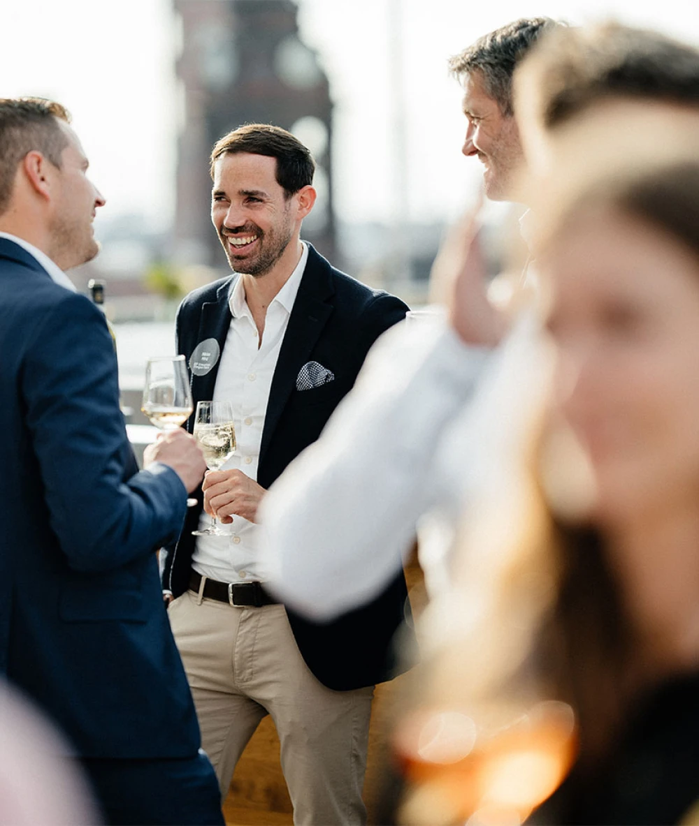 Drei Männer unterhalten sich lächelnd bei einem gesellschaftlichen Anlass mit Weingläsern in der Hand
