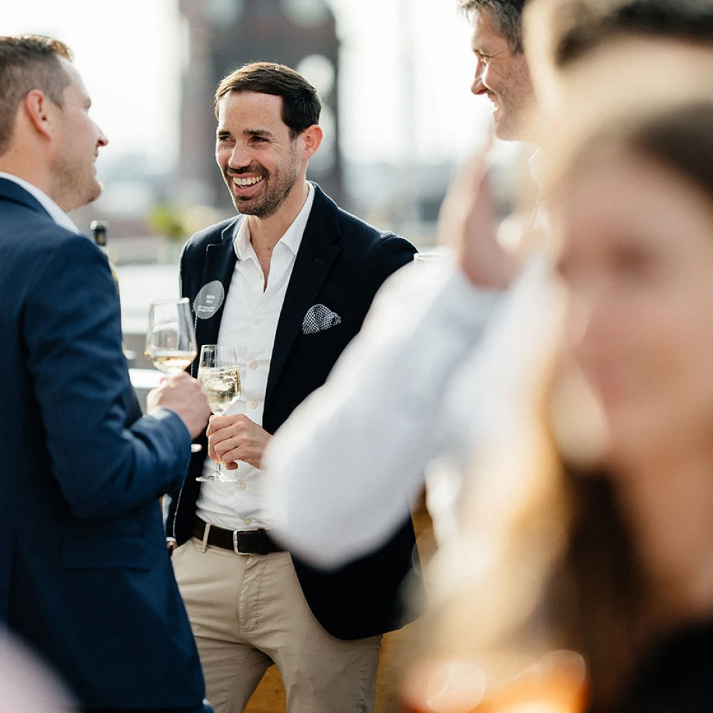 Drei Männer unterhalten sich lächelnd bei einem gesellschaftlichen Anlass mit Weingläsern in der Hand
