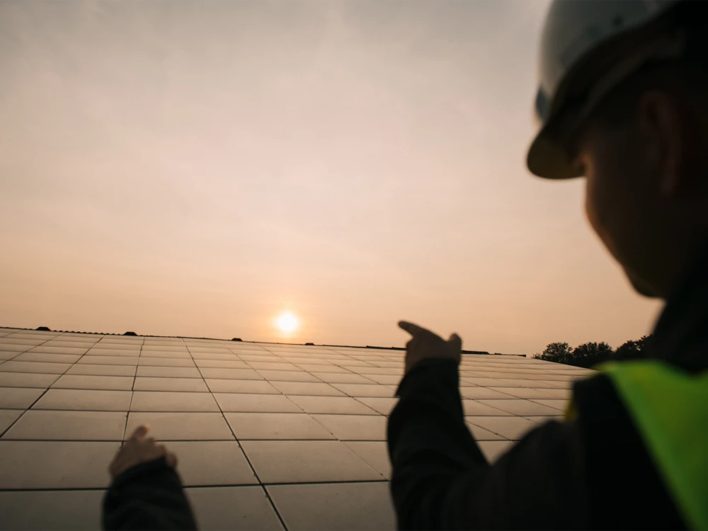 Person in Arbeitskleidung und Helm deutet auf Solarmodule bei Sonnenuntergang