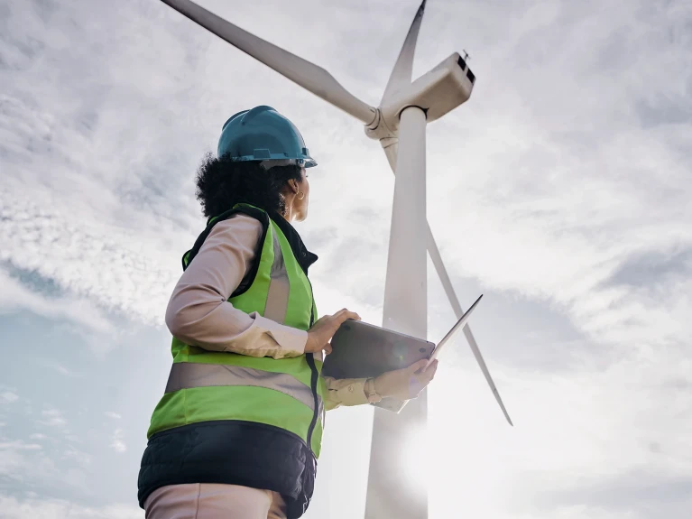 Engineer wearing helmet and reflective vest holding a clipboard and looking at a wind turbine