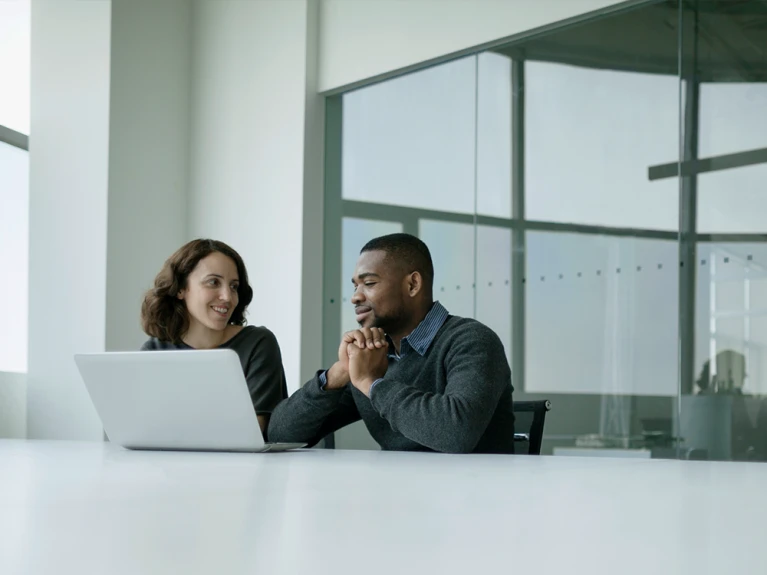 Two business professionals, a woman and a man, sitting at a table looking at a laptop in a bright office