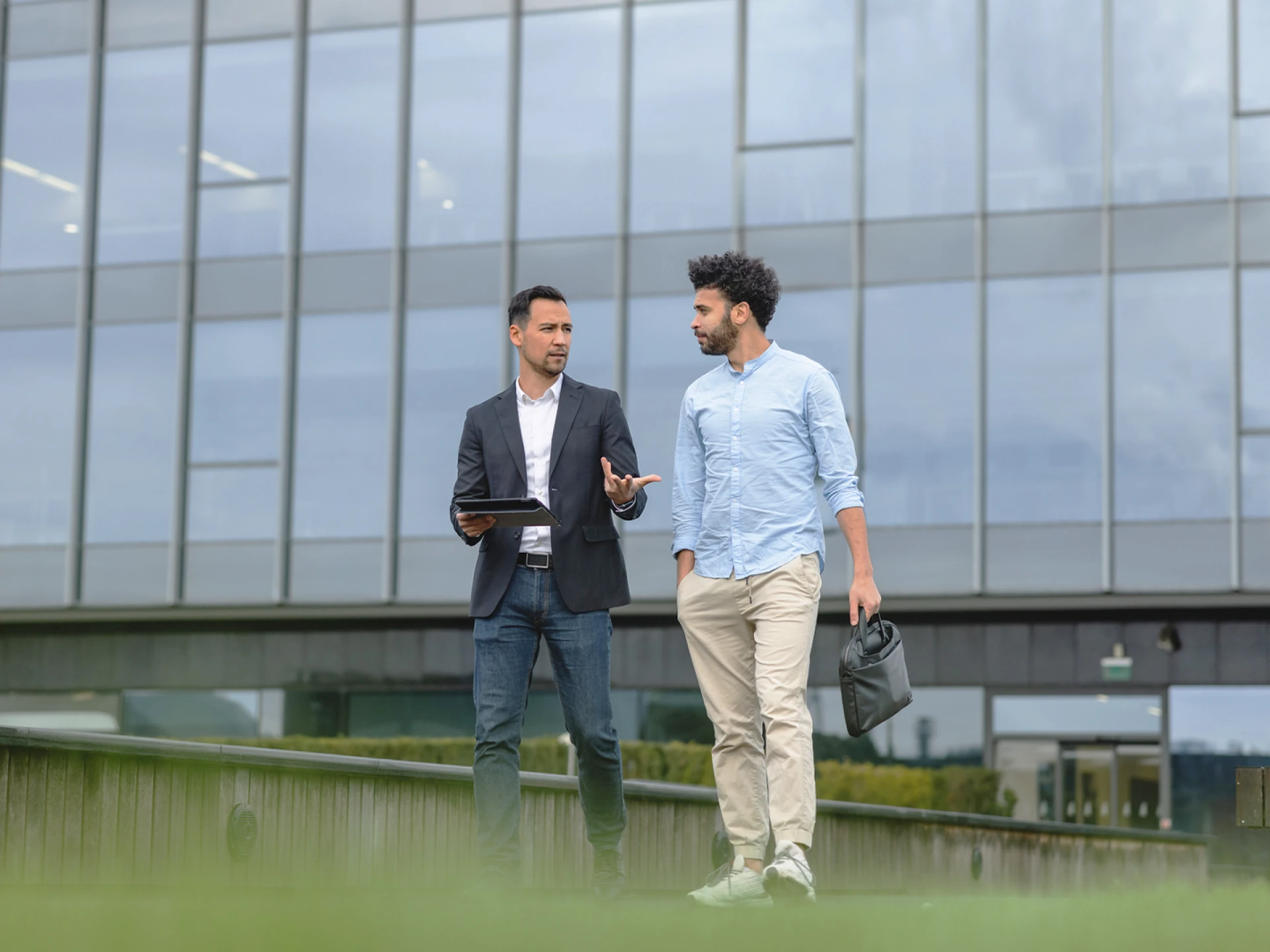 Two men talking while walking in front of a modern building with a glass facade