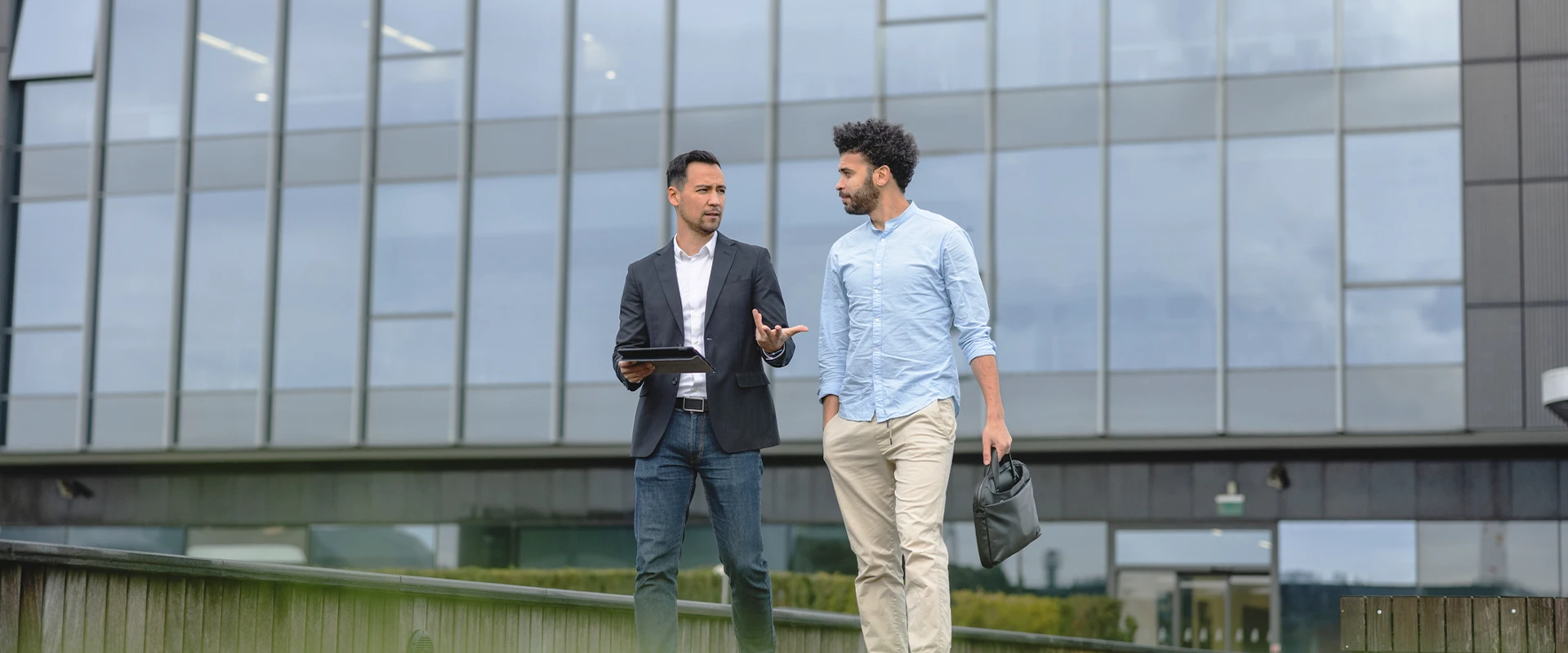 Two men talking while walking in front of a modern building with a glass facade