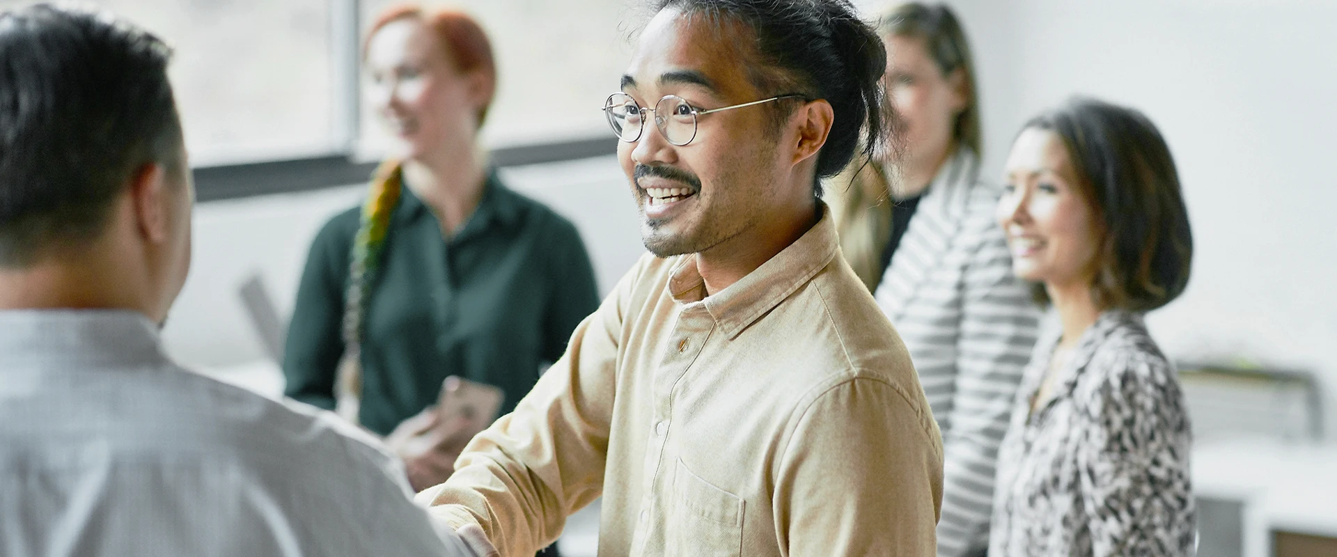 A man with glasses and a shirt shaking hands with another man in an office with colleagues in the background