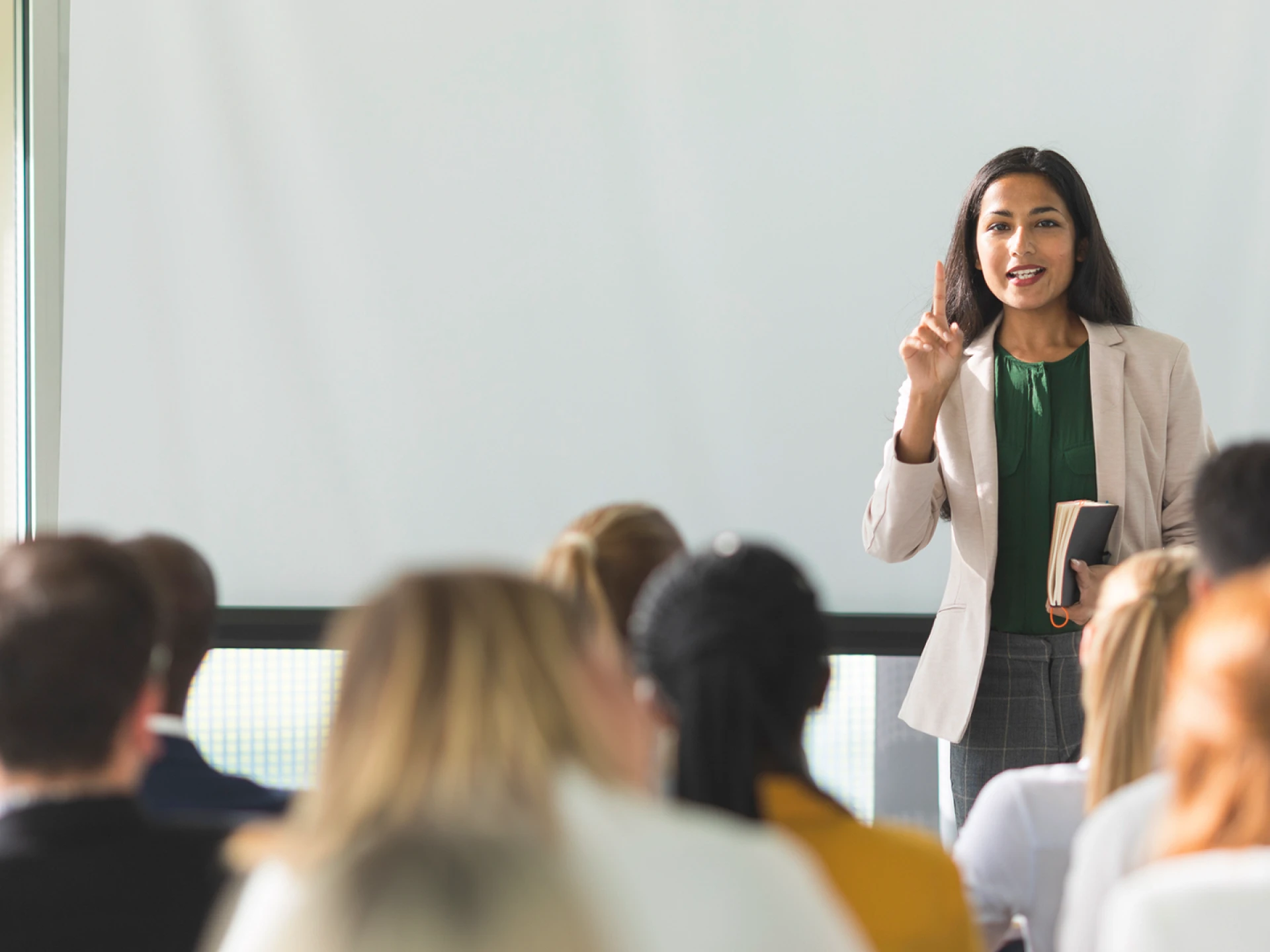 A woman stands in front of a group of listeners and speaks during a presentation.