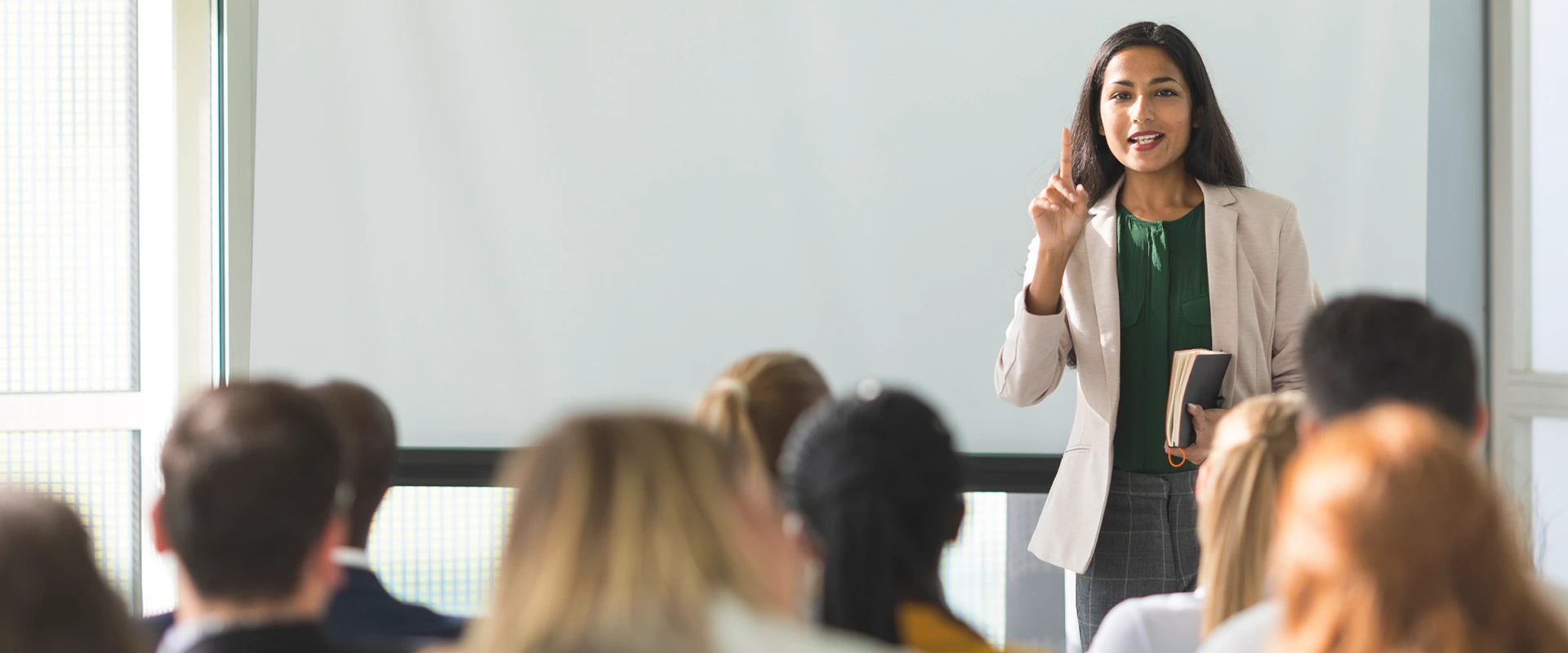 A woman stands in front of a group of listeners and speaks during a presentation.