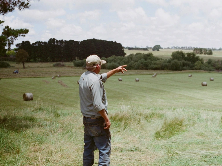Elderly man with cap pointing over a field with hay bales