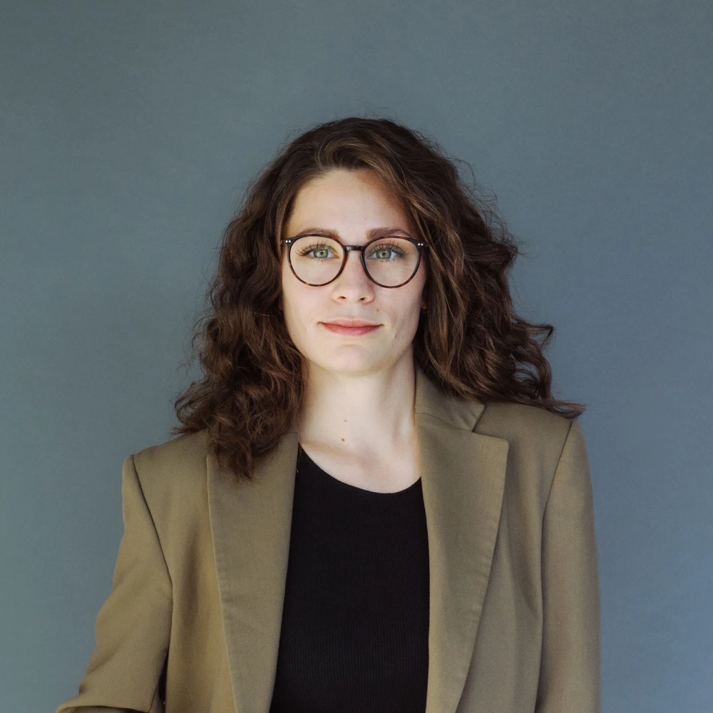 Porträt einer jungen Frau mit Brille und lockigem Haar vor einem grauen Hintergrund