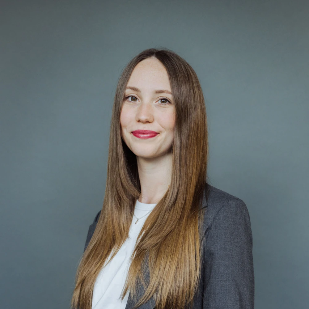 Porträt einer jungen Frau mit langen glatten Haaren und einem grauen Blazer vor einem grauen Hintergrund