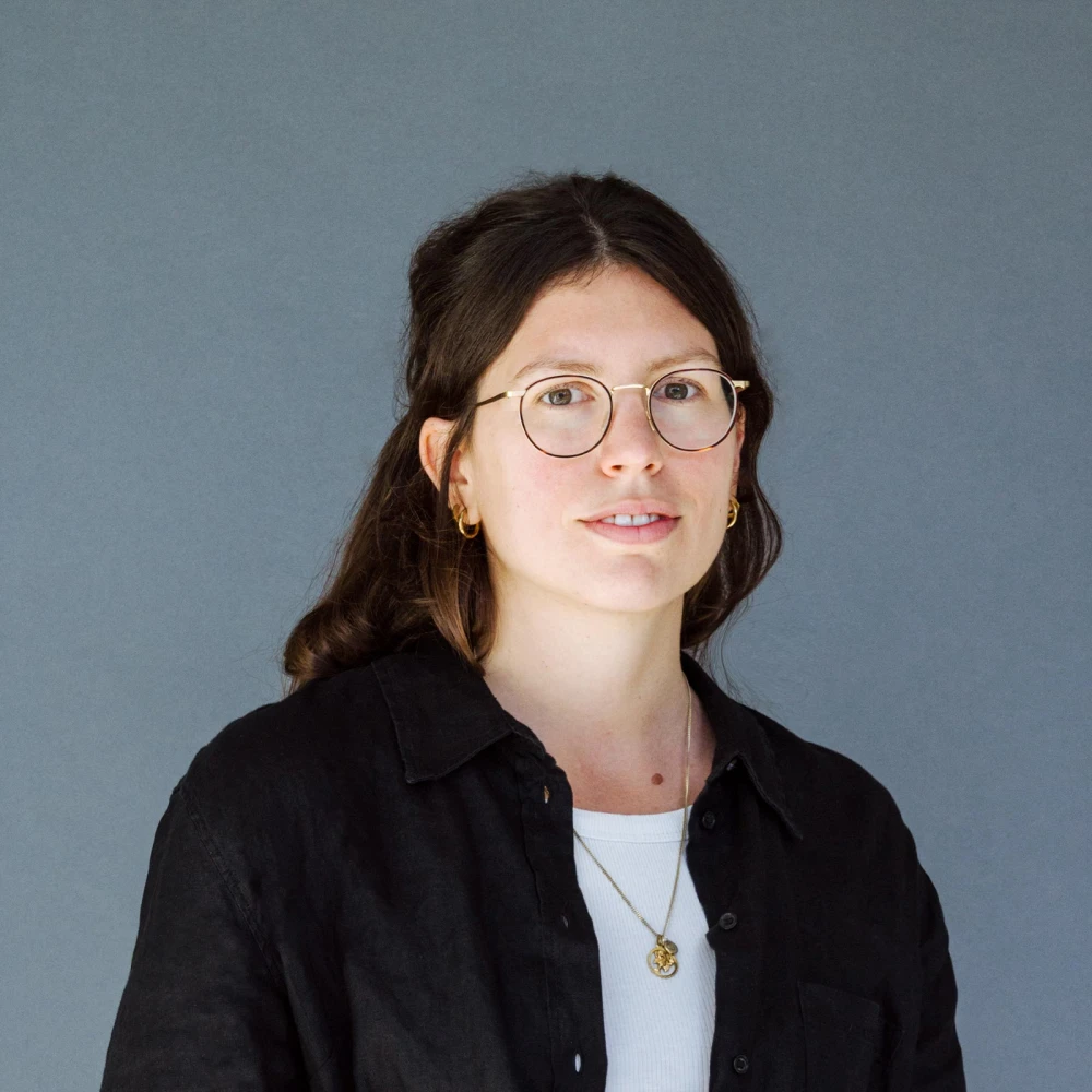 Porträt einer jungen Frau mit Brille und schwarzer Bluse vor grauem Hintergrund