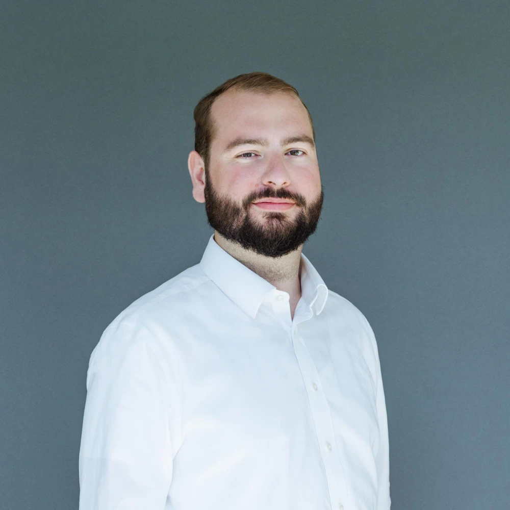 Porträt eines lächelnden Mannes mit Bart in weißem Hemd vor grauem Hintergrund