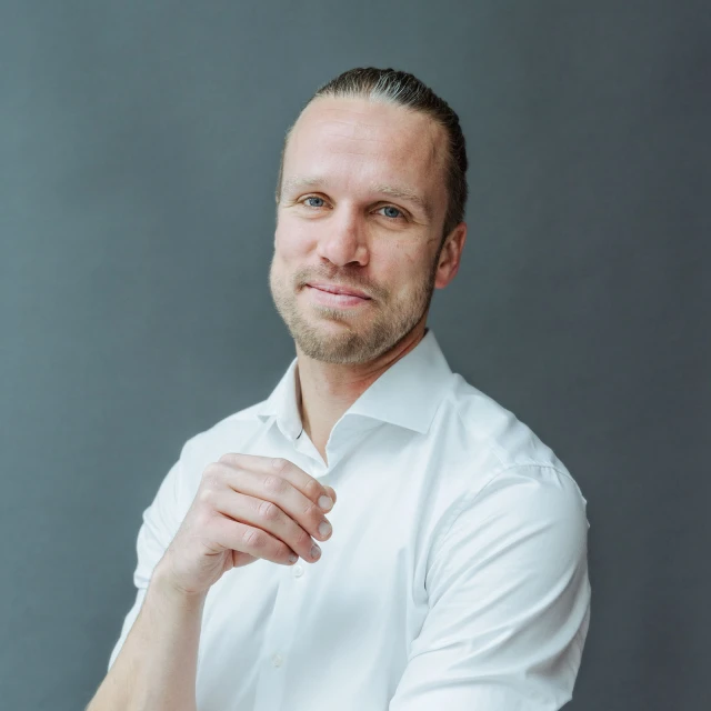 Portrait of a smiling man with slicked-back blonde hair wearing a white shirt against a gray background