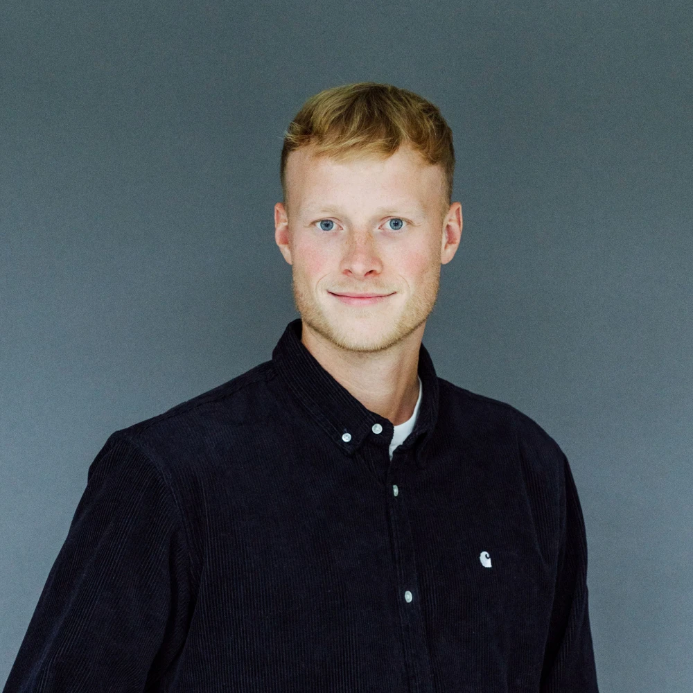 Porträt eines lächelnden jungen Mannes mit roten Haaren und einem dunkelblauen Hemd vor einem grauen Hintergrund