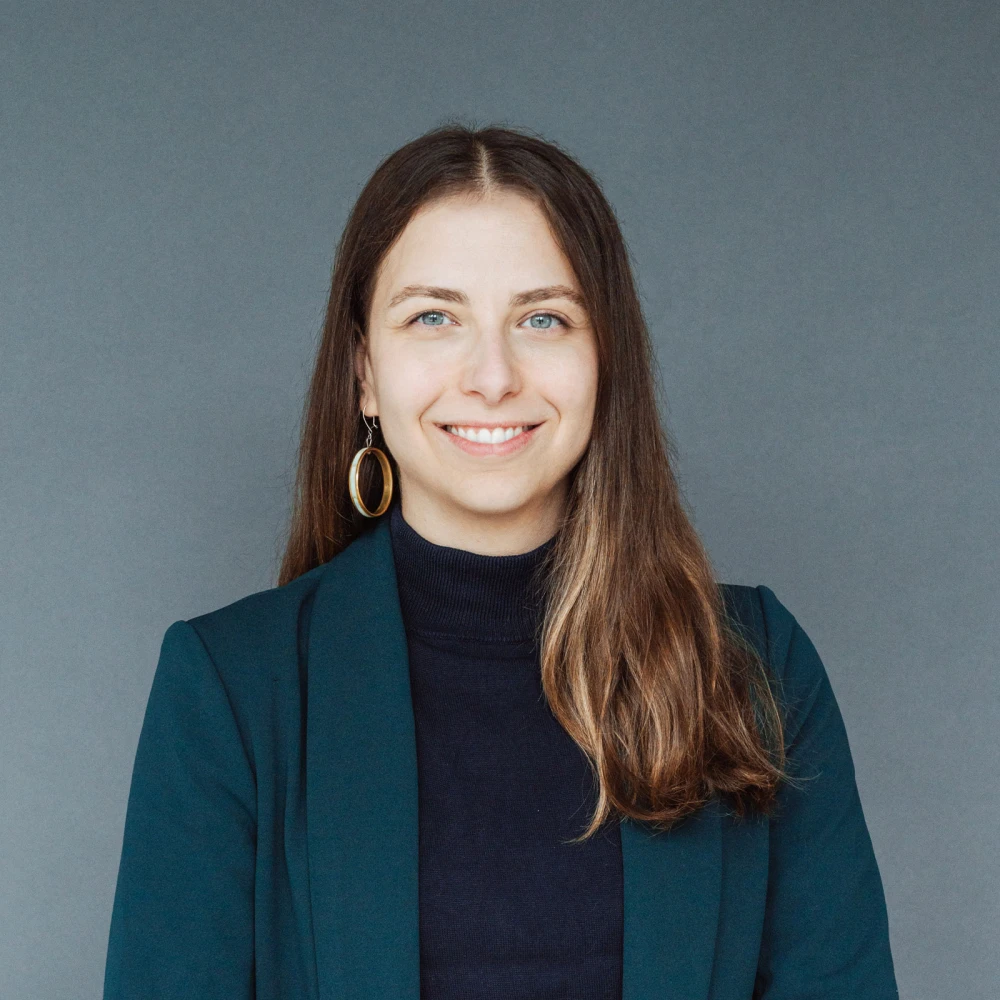 Porträt einer lächelnden Frau mit langen braunen Haaren, blauen Augen und einem dunkelblauen Rollkragenpullover unter einem tealgrünen Blazer.