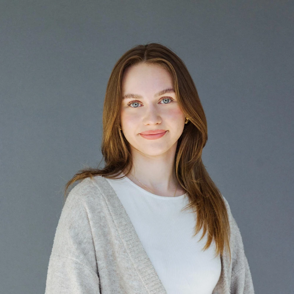 Portrait of a young woman with long brown hair and a white top against a gray background