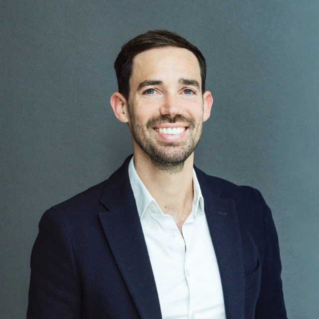 Porträt eines lächelnden Mannes mit dunklem Haar und Bart, der ein dunkelblaues Sakko und ein weißes Hemd trägt