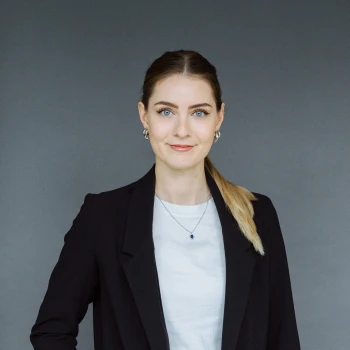 Portrait of a young woman with tied-back blonde hair wearing a black jacket and white shirt, against a grey background