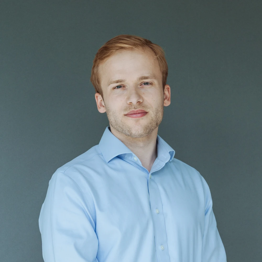 Portrait of a smiling man with light blonde hair and blue shirt against a grey background