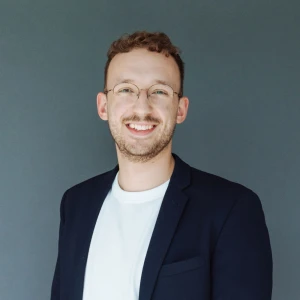 Lächelnder junger Mann mit Brille und Bart, trägt ein weißes T-Shirt und einen dunkelblauen Blazer vor einem grauen Hintergrund