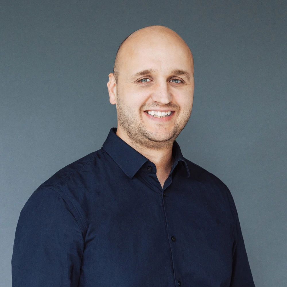 Porträt eines lächelnden kahlen Mannes in dunkelblauem Hemd vor grauem Hintergrund