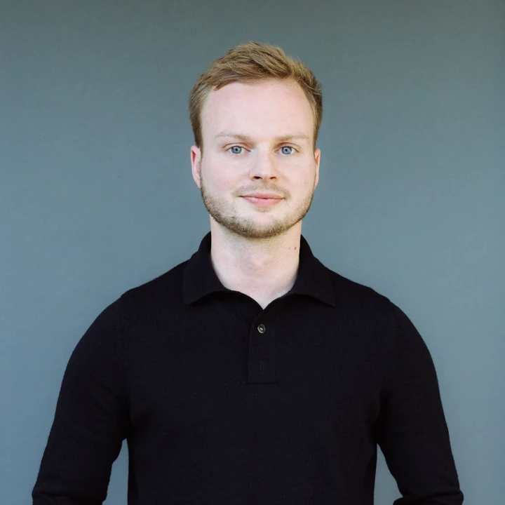 Porträt eines jungen Mannes mit blonden Haaren und einem schwarzen Poloshirt vor einem grauen Hintergrund