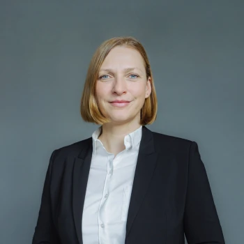 Portrait of a smiling woman with medium-length blonde hair wearing a black suit and white shirt against a gray background