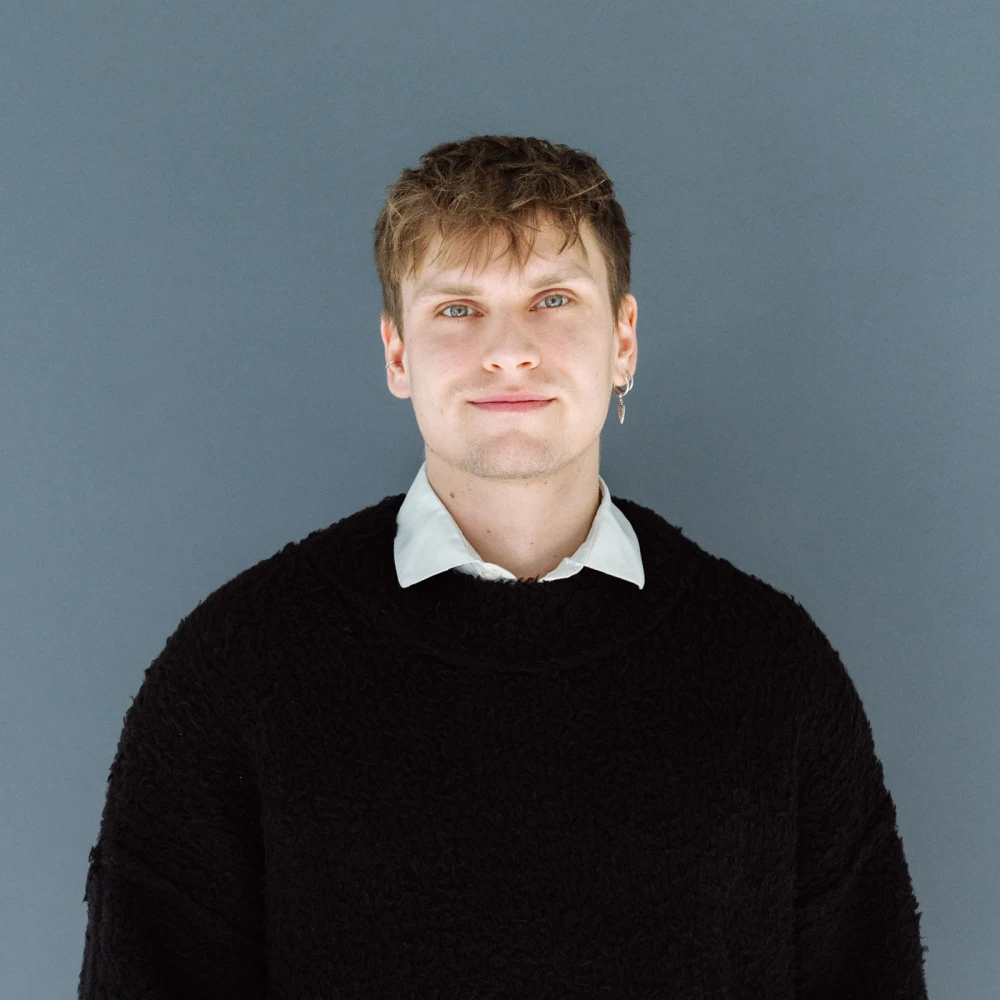 Porträt eines jungen Mannes mit blonden Haaren und einem Ohrring, der einen schwarzen Pullover und ein weißes Hemd trägt